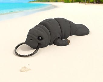 May include: A dark gray manatee keychain with a black keyring. The manatee is lying on a sandy beach with a turquoise ocean and palm trees in the background. The manatee is a detailed, three-dimensional design.
