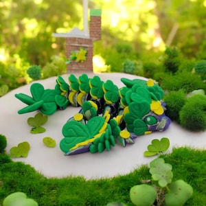 May include: A green and yellow clover-shaped dragon figurine with purple accents. The dragon is lying on a white surface with green moss around it.