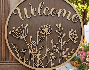 May include: A round wooden sign with the word "Welcome" in a decorative font. The sign is painted brown and features a laser-cut design of stylized flowers and leaves in a gold color.
