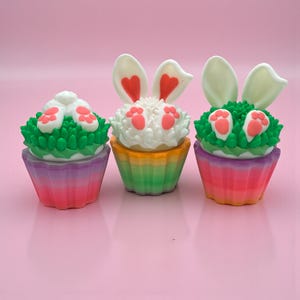 Puede incluir: Tres cupcakes con temática de Pascua con orejas y patas de conejo. Los cupcakes tienen glaseado colorido y están decorados con glaseado blanco, glaseado verde y detalles en rosa. Los moldes para cupcakes son en tonos rosa, morado, naranja y verde.