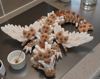 May include: A whimsical dragon sculpture crafted from cookies and cream elements. The dragon features detailed wings, a cookie-textured body, and a head with cookie accents. The sculpture is displayed on a metallic surface.