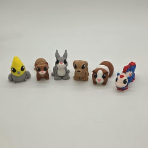 Peut inclure: Ensemble de six figurines d'animaux miniatures. L'ensemble comprend un oiseau jaune et gris, un écureuil brun, un lapin gris, un chien beige, un cochon d'Inde brun et blanc et un poisson rouge, blanc et bleu.