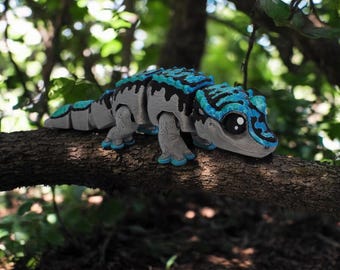 May include: A blue and black toy lizard with large eyes, perched on a tree branch. The lizard has a textured, segmented body and is made of plastic.
