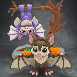 May include: Two bat figurines featuring one brown bat standing on a gray base with two orange pumpkins and one purple bat hanging upside down from a brown tree branch. Both bats have large eyes, pink noses, and cream-colored chest fur.
