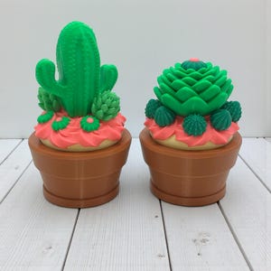 May include: Two decorative cactus sculptures in brown pots. One cactus is tall and green, the other is a layered green design. Both have pink and green accents, resembling frosting and flowers. The pots are a warm brown color.