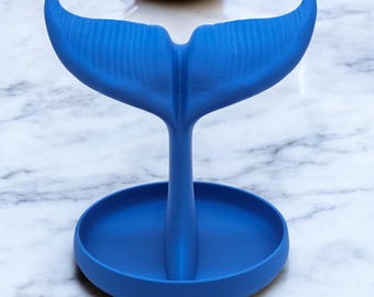 May include: A blue whale tail shaped ring holder with a round base. The holder is made of a smooth, plastic material.