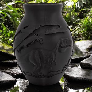 Puede incluir: Un jarrón de cerámica negro con un diseño de dinosaurio. El jarrón es redondo y tiene una boca ancha. El dinosaurio está en relieve y corre por un paisaje de bosque.