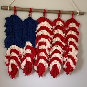 May include: A macrame wall hanging featuring five feather-shaped fiber art pieces suspended from a horizontal wooden dowel. The fibers are knotted in red, white, and blue, arranged to resemble the pattern of the American flag with a hanging cord.