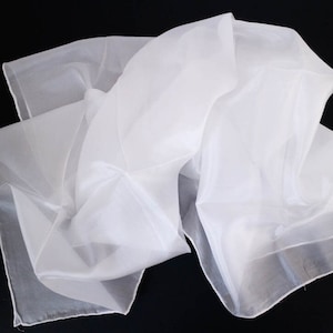 Peut inclure: Une écharpe blanche en voile avec un léger reflet irisé. L'écharpe est pliée et drapée sur un fond noir.