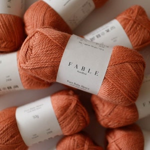 Peut inclure: Gros plan sur plusieurs écheveaux de fil orange étiquetés "FABLE handknit Pure Baby Alpaca 100% Baby Alpaca 50g environ 145 yds / 132 m"