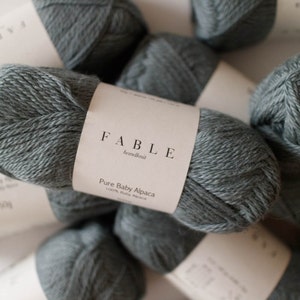 May include: A collection of grey Fable handknit Pure Baby Alpaca yarn skeins. Each ball has a white label with the brand name and yarn details. The yarn is a soft, muted grey colour, and the image is well-lit.
