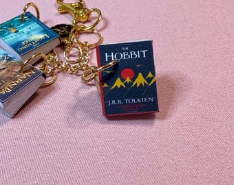 May include: Miniature book keychains featuring titles like "The Hobbit" and "The Lightning Thief." The keychains have gold-colored hardware, including rings, a chain, and a clasp. The books have colorful covers with text and illustrations.