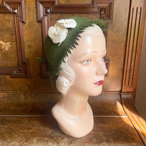Peut inclure: Un chapeau vert vintage avec des accents floraux blancs est présenté sur une tête de mannequin. Le chapeau présente un design unique avec un bord texturé et de délicates fleurs blanches. La tête de mannequin est posée sur une surface en bois.