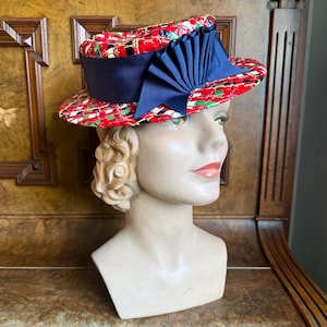 Puede incluir: Un sombrero vintage con una corona estampada en rojo, verde y negro y una cinta ancha azul marino. El sombrero se exhibe en una cabeza de maniquí. El ala del sombrero está ligeramente levantada y la cinta azul marino está atada en un gran lazo.