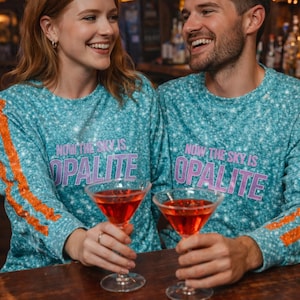 May include: A couple smiles while holding martini glasses. They wear matching teal long-sleeve shirts with the text "NOW THE SKY IS OPALITE" and orange stripes on the sleeves. The drinks are red, and the background is a bar.