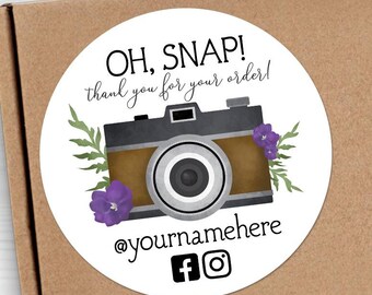 May include: Round sticker with a white background, featuring a cartoon illustration of a vintage camera with purple flowers and green leaves. The text "OH, SNAP! thank you for your order!" is written in black cursive font. Below the camera, the text "@yournamehere" is written in black font, followed by Facebook and Instagram icons.