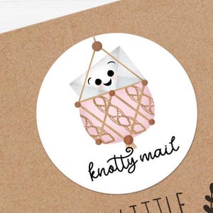 May include: A white envelope with a smiling face is inside a pink and brown macrame hanging. The text "knotty mail" is written below the hanging.