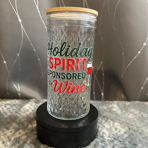 May include: A clear glass jar with a diamond pattern, a wooden lid, and the text "Holiday Spirit Sponsored by Wine" in green and red. A wine glass graphic is also included. The jar is on a black stand.