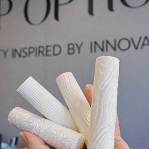 Könnte beinhalten: Eine Hand hält vier weiße, zylindrische Objekte mit strukturierten Mustern. Die Muster umfassen wellenförmige Linien, vertikale Rippen und gepunktete Designs. Im Hintergrund stehen die Worte "P OPTIC" und "TY INSPIRED BY INNOVA".