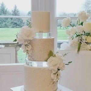 May include: A four-tiered wedding cake on a white pedestal. The top tier is a plain, light-colored cylinder. The second tier is covered in sparkling embellishments. The bottom two tiers are white with floral designs and decorative trim. White flowers cascade down the sides.