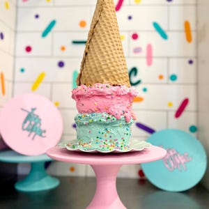 May include: A layered cake with pink and light blue frosting, topped with a large waffle cone. The cake is decorated with colorful sprinkles and sits on a light pink pedestal stand. The background features a white tiled wall with colorful sprinkles.