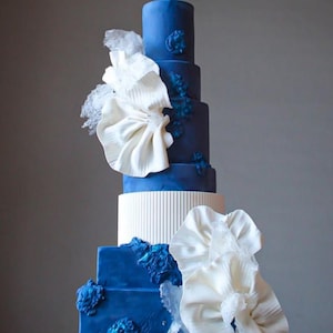 Könnte beinhalten: Ein mehrstöckiger Kuchen mit auffälligem Design. Der Kuchen hat Schichten in tiefem Blau und Weiß, verziert mit großen, gerüschten weißen Akzenten und blauen floralen Verzierungen. Der Kuchen hat eine quadratische und runde Form.