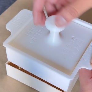 Könnte beinhalten: Ein weißes Tortenwerkzeug aus Kunststoff mit einem zentralen Griff und seitlichen Laschen, das den geprägten Text "PROP OPTIONS" und "KEEP CAKE" aufweist. Das Werkzeug wird auf eine rechteckige Basis gedrückt, wobei der Text "CAKE PALETTES Using a custom cutter" über das Bild gelegt ist.