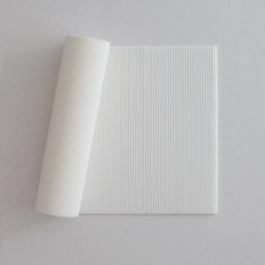May include: A white, textured, rectangular mat with vertical ridges, partially rolled up. The mat is made of a ridged material and is unrolled to reveal its full rectangular shape. The background is a light, speckled surface.
