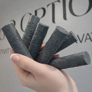 May include: A set of five cylindrical dark grey rollers, each featuring a distinct embossed pattern including floral motifs, intricate lace-like textures, and wood grain designs, held together in a fan-like arrangement.