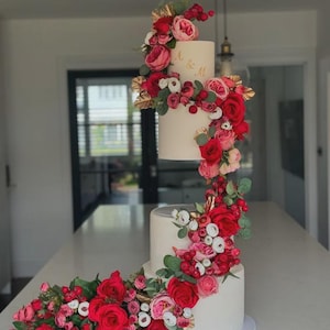 Könnte beinhalten: Ein dreistöckiger weißer Kuchen, verziert mit roten und rosa Rosen, Beeren und goldenen Akzenten. Die oberste Etage hat goldene Buchstaben. Ein wunderschöner Kuchen für eine Hochzeit oder einen besonderen Anlass.