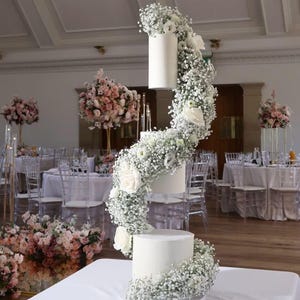 Könnte beinhalten: Eine dreistöckige weiße Hochzeitstorte, die mit weißen Gipskrautblüten verziert ist, die an den Seiten herunterfallen. Die Torte steht auf einem weißen Tisch in einem Raum mit weißen Wänden und durchsichtigen Stühlen.