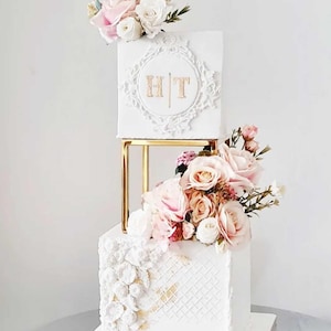 Może przedstawiać: Trzywarstwowy biały tort ślubny z złotymi akcentami. Górny poziom to biały kwadratowy tort z złotym monogramem "HT" w ozdobnej ramie. Środkowy poziom to złoty kwadratowy stojak z białym kwadratowym tortem. Dolny poziom to biały kwadratowy tort z białym wzorem koronkowym i złotymi akcentami. Tort jest ozdobiony różowymi i białymi kwiatami.