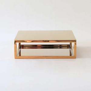 Op de afbeelding: Een gouden metalen tweelaags standaard met een rechthoekige vorm. De standaard heeft een gespiegelde oppervlak.