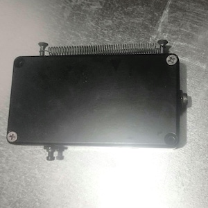 Può includere: Un dispositivo rettangolare nero con una molla e viti attaccate. Il dispositivo ha una superficie liscia e sembra essere in metallo. Le viti sono argentate e la molla è avvolta. Il dispositivo è su una superficie grigia.