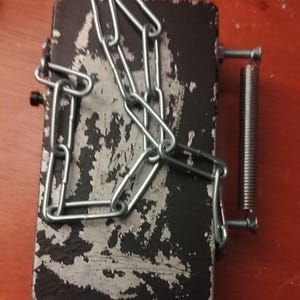 Könnte beinhalten: Eine schwarze Metallbox mit einer silbernen Kette und einer daran befestigten Feder. Die Box hat ein abgenutztes und verwittertes Aussehen.
