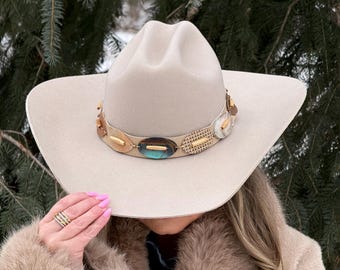 Könnte beinhalten: Ein hellbeiger Filz-Cowboyhut mit einem dekorativen Hutband, das mit verschiedenen ovalen Anhängern verziert ist, darunter goldfarbene, schillernd blaue und texturierte beige Elemente, die den unteren Teil der Krone umschließen.