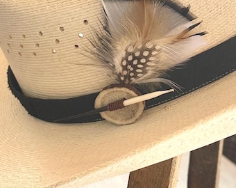Op de afbeelding: Een strooien hoed met een zwarte band en decoratieve veren. De veren zijn bruin, wit en zwart, bevestigd aan een kleine, ronde houten knoop met een botachtig accent. De hoed heeft kleine, gelijkmatig verdeelde gaatjes.