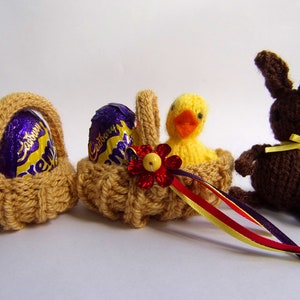 Könnte beinhalten: Drei gestrickte Osterdekorationen: zwei Körbe mit Cadbury Creme Eggs, ein gelbes Küken in einem Korb und ein braunes Häschen mit gelber Schleife. Die Körbe haben rote und gelbe Blumenakzente.