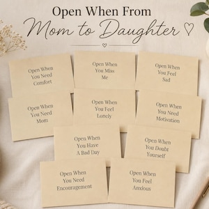 May include: A collection of beige cards with handwritten text, arranged on a neutral surface. The cards read "Open When" followed by various emotional prompts, such as "You Need Comfort" and "You Feel Sad." The phrase "Open When From Mom to Daughter" is at the top.