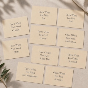 May include: A collection of beige cards with black text, arranged on a neutral surface. Each card reads "Open When" followed by a specific emotion or need, such as "Comfort," "Sad," or "Motivation." A sprig of greenery is visible.