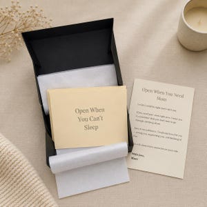 May include: A black gift box with a stack of cream-colored cards, one labeled "Open When You Can't Sleep." A separate card reads "Open When You Need Mom" with a handwritten message. The box is open, revealing tissue paper.
