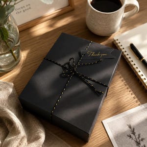 May include: A black gift box tied with black and gold twine, with a small "Thank you" tag. A mug of coffee, a notebook, and a pen are on a wooden surface. A framed picture and a vase with greenery are also present.