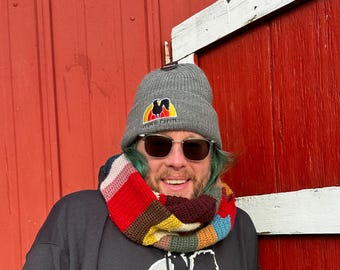 Puede incluir: Un gorro gris con el logo "Dawn Farm", usado con un cuello de ganchillo colorido y gafas de sol. El cuello presenta un diseño de patchwork con secciones rojas, marrones, amarillas y azules. La persona lleva una sudadera con capucha negra.