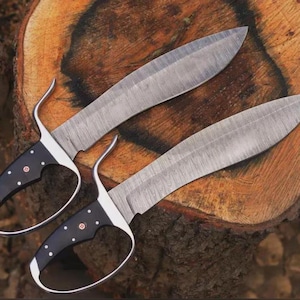 May include: Two Damascus steel knives with black handles and finger loops. The blades have a curved shape and are resting on a wooden surface. The handles are black with white dot accents and a red emblem.