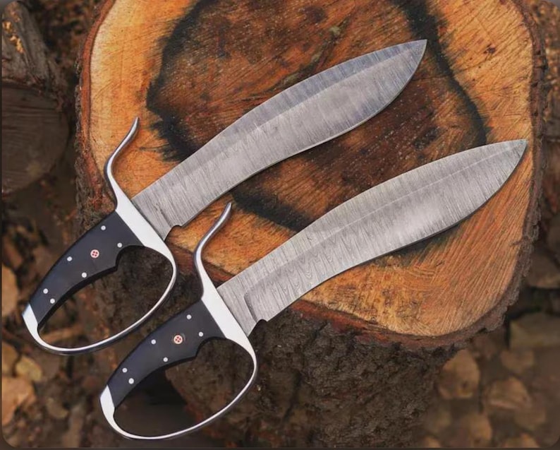 May include: Two Damascus steel knives with black handles and finger loops. The blades have a curved shape and are resting on a wooden surface. The handles are black with white dot accents and a red emblem.