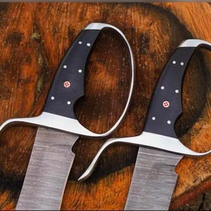 May include: Two swords with curved silver handles and black grips adorned with white dots and a red emblem. The blades have a textured, gray appearance. The swords are displayed on a wooden surface.