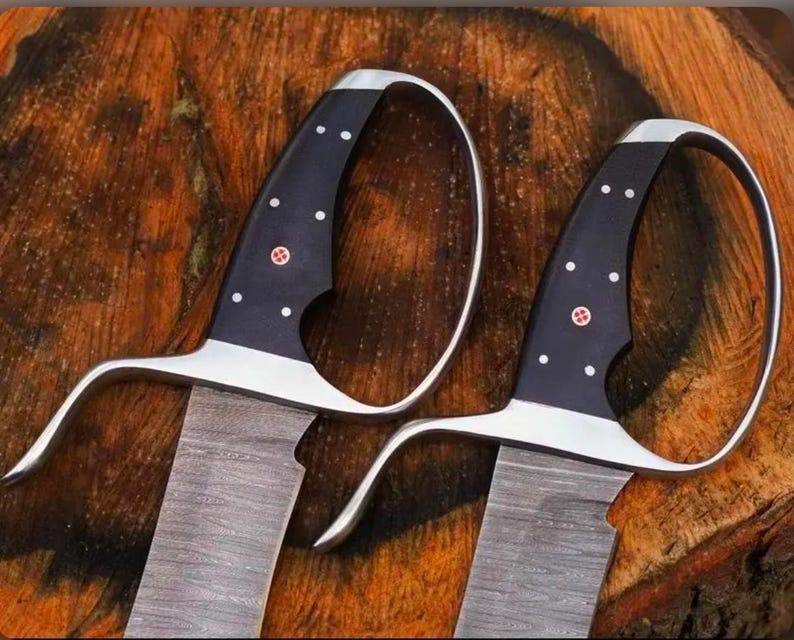 May include: Two swords with curved silver handles and black grips adorned with white dots and a red emblem. The blades have a textured, gray appearance. The swords are displayed on a wooden surface.
