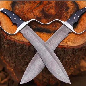 May include: Two crossed, hand-forged knives with curved handles and wide blades. The handles are black with white dot accents and a silver guard. The blades are gray with a textured pattern. The knives are displayed on a wooden tree trunk.