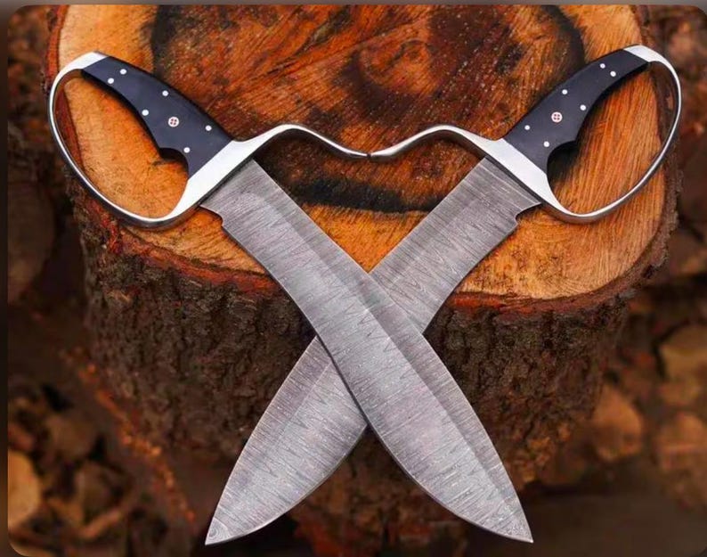 May include: Two crossed, hand-forged knives with curved handles and wide blades. The handles are black with white dot accents and a silver guard. The blades are gray with a textured pattern. The knives are displayed on a wooden tree trunk.