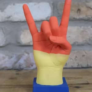May include: A hand sculpture in a rock-on gesture, painted in orange, yellow, and blue. The fingers are orange, the palm is yellow, and the base is blue. The sculpture is on a wooden surface.